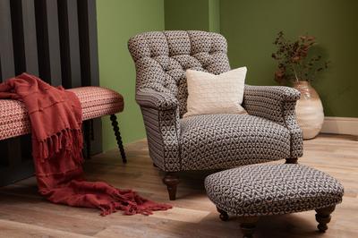 A hand-buttoned Slipper Armchair and ottoman in patterned dark fabric, Emilys Maze Midnight, with a cream pillow against a green wall.