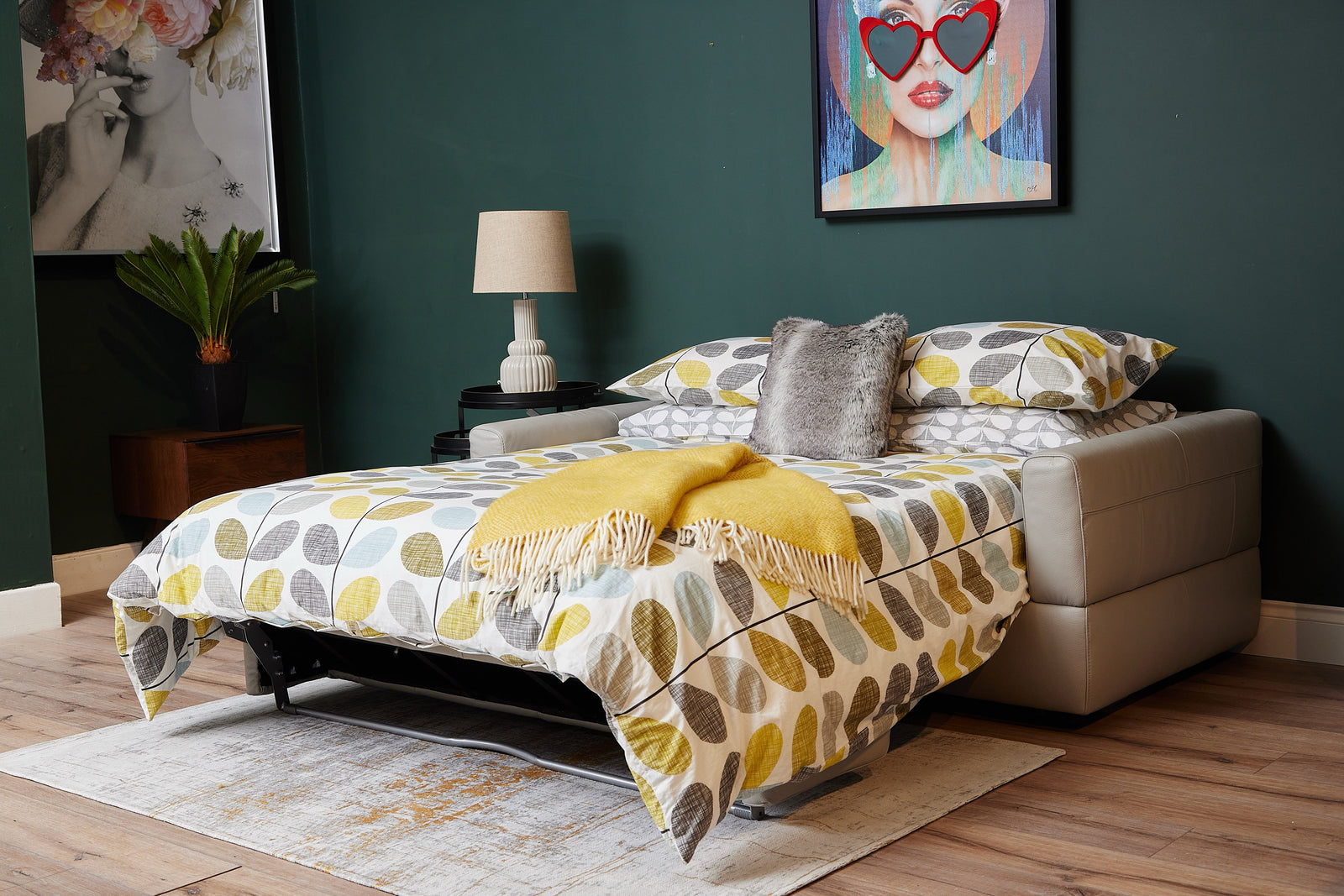 Modern sofa bed extended into a full bed with patterned bedding, a yellow throw, and decorative pillows in a stylish room with artwork and warm wood flooring.