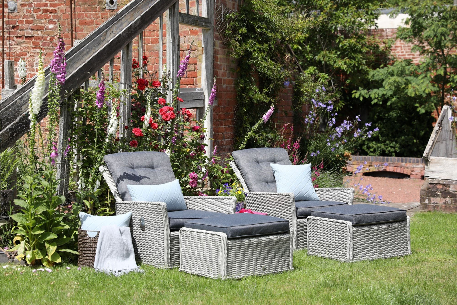Two grey wicker garden lounge chairs with matching footstools and cushions, arranged on a lawn beside colourful flowers and a rustic brick wall.
