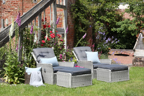 Two grey wicker garden lounge chairs with matching footstools and cushions, arranged on a lawn beside colourful flowers and a rustic brick wall.