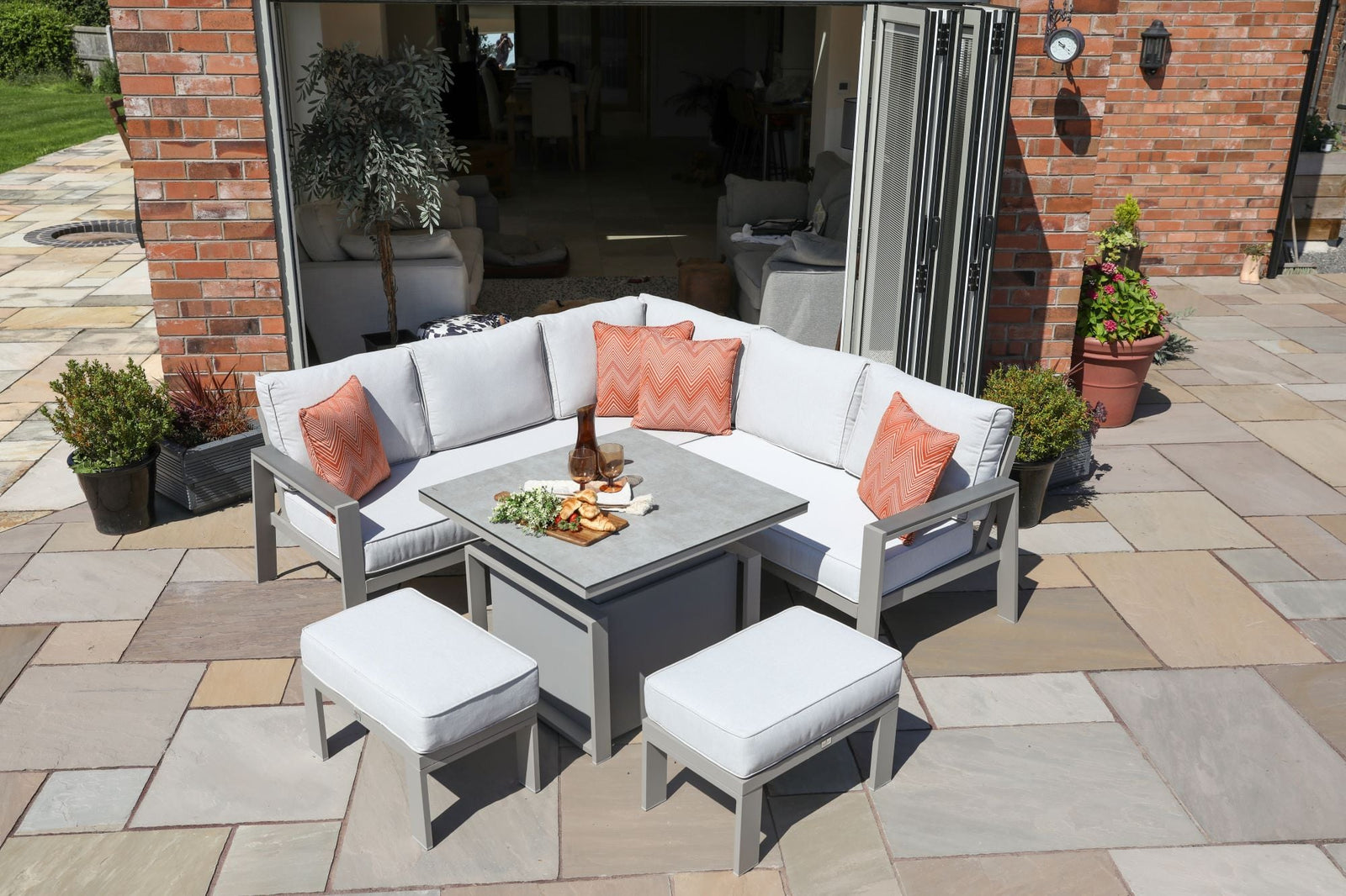 A white-cushioned gray outdoor sectional with orange throw pillows surrounds a square coffee table on a stone-paver patio outside a brick home with open folding doors.