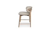 A modern counter stool with a light beige upholstered seat and curved backrest, supported by a natural wood frame with integrated armrests and slim legs.