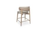 A modern bar stool with a curved light beige upholstered seat and backrest, supported by a natural oak wood frame with a square crossbar.