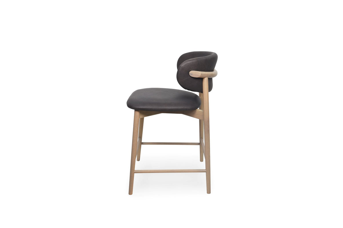A modern bar stool with a curved chocolate upholstered backrest and seat, supported by a light natural wood frame with a curved wooden armrest and four legs, shown on a solid white background.