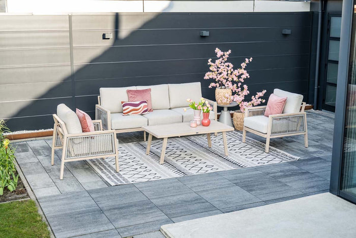 Neutral outdoor lounge set with cream sofa and armchairs, pink cushions, and a low coffee table on a patterned rug in a modern paved patio.