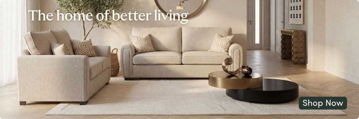 A staged living room scene showing a light beige sofa and matching armchair with decorative pillows, a round coffee table, and a rug. Large text at the top reads 'The home of better living' and there is a 'Shop Now' button in the lower right corner.
