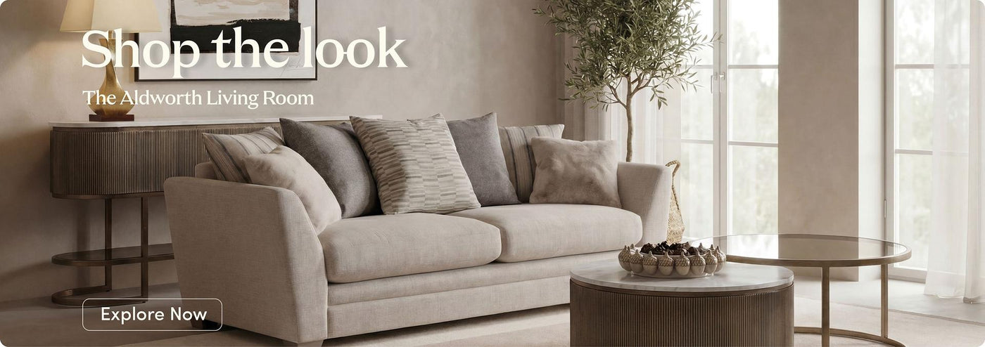 A beige upholstered sofa with multiple gray and taupe throw pillows in a bright, modern living room. Large windows, a side table with a lamp, and a round coffee table are visible.