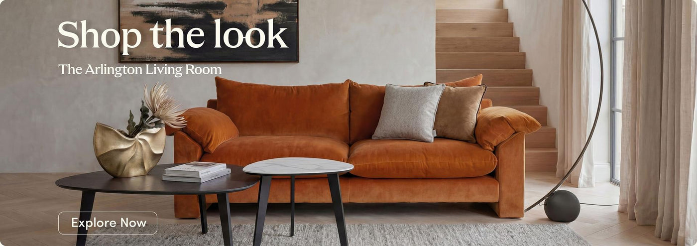 An orange-brown velvet sofa in a styled living room setting, featuring a black coffee table, a round white-top side table, a curved floor lamp, and a staircase in the background. Text overlays read 'Shop the look', 'The Arlington Living Room', and 'Explore Now'.
