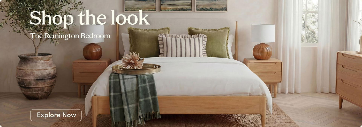 A styled bedroom scene featuring a light wood bed frame with white bedding, green and striped pillows, matching nightstands, lamps, a dresser, and a large vase with a plant. Text on the image reads 'Shop the look The Remington Bedroom' with an 'Explore Now' button.
